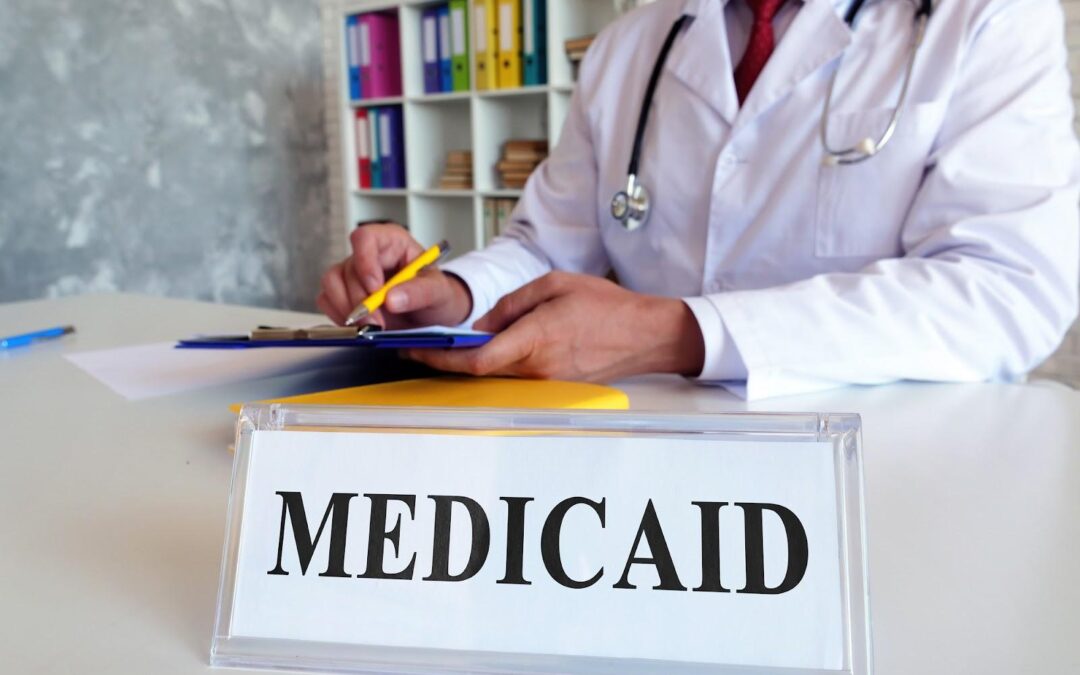 A doctor in a white coat fills out forms for Medicaid planning behind a desk plate that reads “Medicaid.”